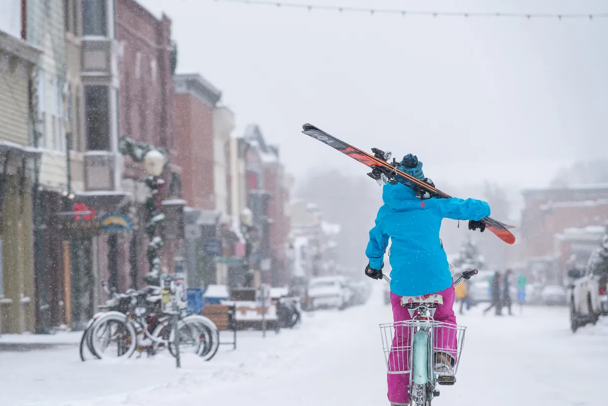 A snow-dusted street bustling with winter activity: a person pedals a bike carrying skis past colorful storefronts, while other bicycles are parked nearby. The lively scene is framed by boutique shop windows and festive decorations.
