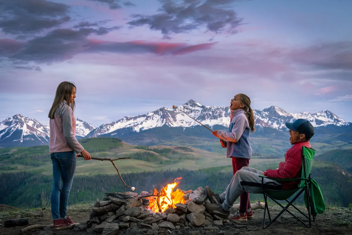A joyful scene of children roasting marshmallows over a campfire set against a majestic mountain backdrop. The environment is lush and natural, emphasizing family bonding, outdoor fun, and the thrill of exploring nature together.