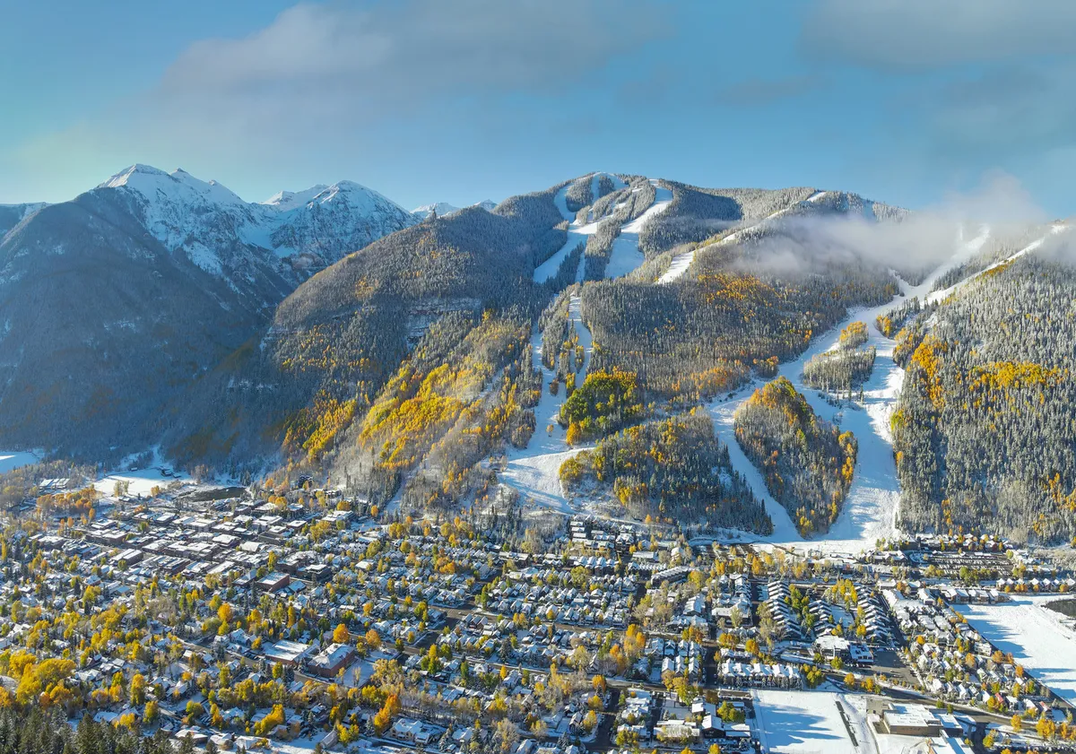 A sweeping view of snow-capped mountains, ski slopes, colorful autumn trees, and the town below. This scene highlights the area's year-round appeal, with vibrant colors suggesting summer and fall hiking and mountain biking as well as winter sports.