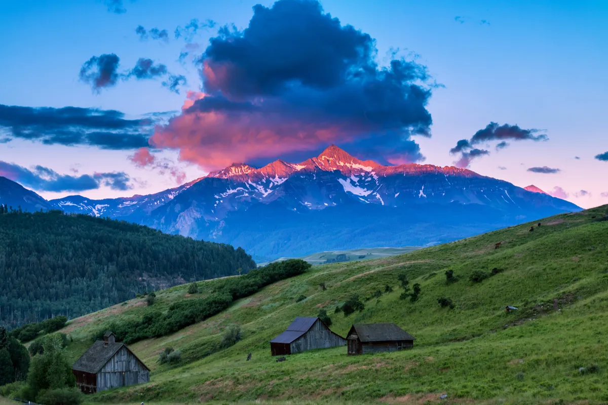 A sweeping view of Telluride’s majestic mountains at sunset, with rustic cabins nestled among lush green hills. The scene evokes a sense of tranquility and natural beauty, and hints at the abundance of nearby hiking trails and outdoor exploration opportunities.
