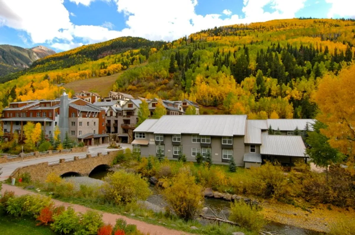 This image captures rolling hills with colorful autumn trees, a modern bridge crossing a gentle stream, and nearby modern buildings. The landscape is tranquil, inviting, and shows open space perfect for strolls, biking, and family picnics. The presence of the bridge and water hints at riverside adventures and child-friendly exploration.