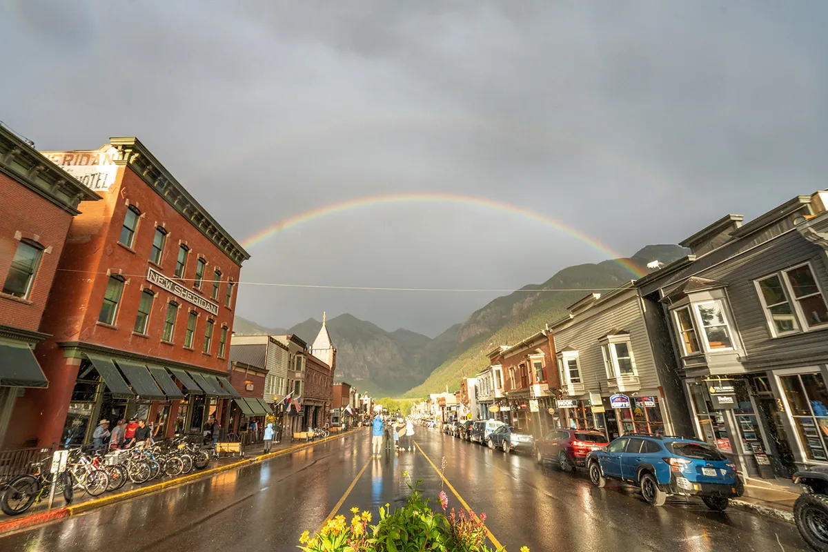 A vibrant street scene featuring historic buildings with a rainbow arching overhead, reflections glistening on the wet street, and people wandering past colorful storefronts. Outdoor cafe tables and bicycles line the sidewalk, evoking a lively, walkable downtown atmosphere.