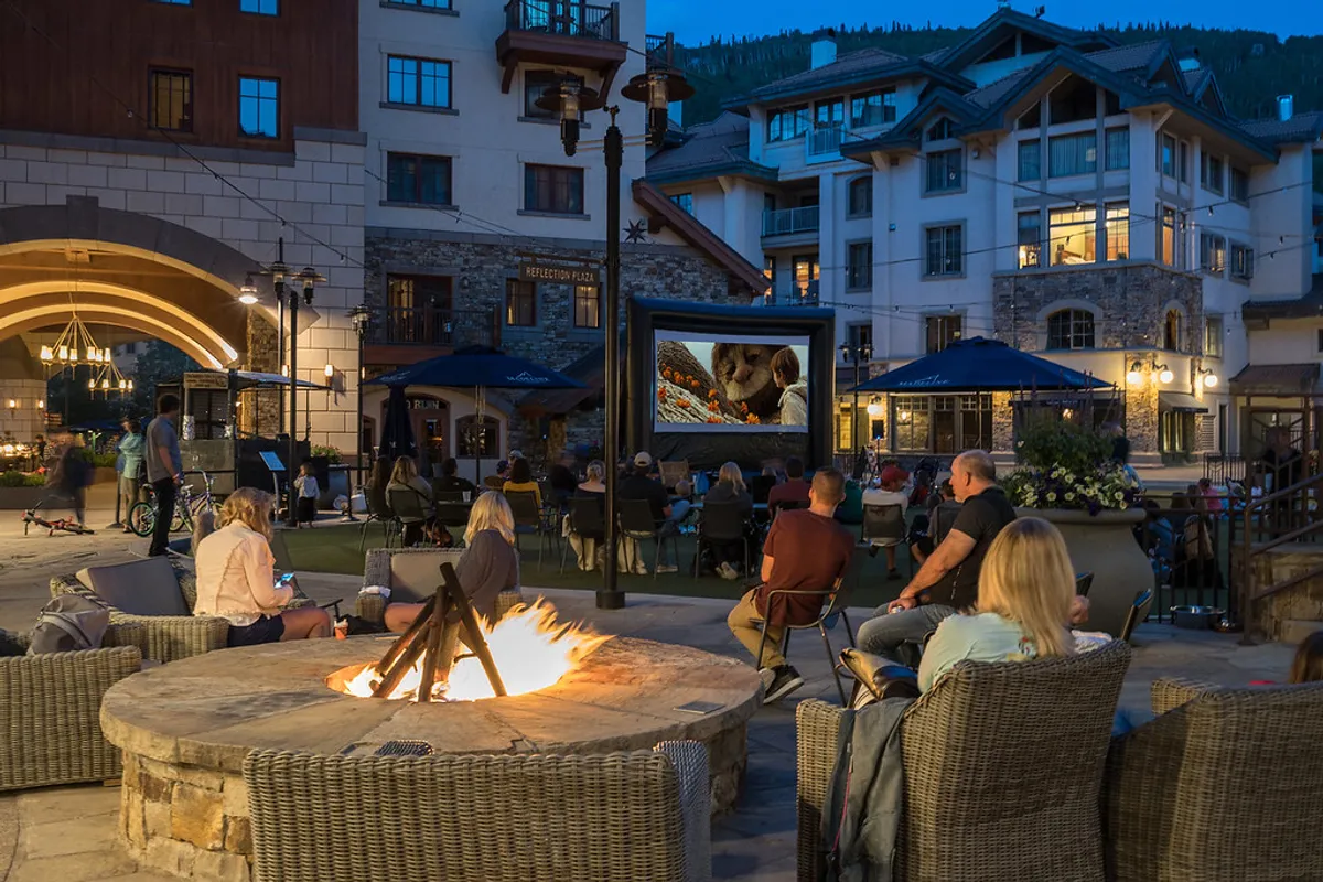 A cozy outdoor gathering space with a glowing fire pit, comfortable seating, and an outdoor movie screen set against the backdrop of Telluride’s architecture. The scene suggests a social, inviting atmosphere ideal for evenings out.