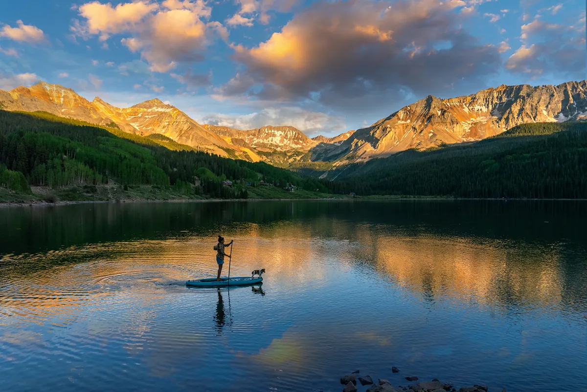 A paddleboarder and their dog glide across a serene lake, reflecting the surrounding mountains. The peaceful scene showcases water-based recreation, nature exploration, and the inclusive, pet-friendly spirit of Telluride summers.