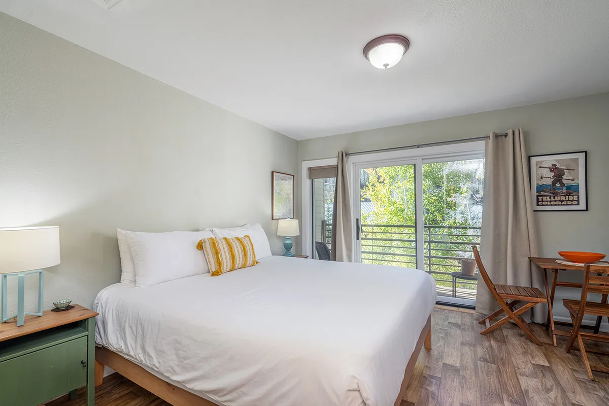 This interior image features a serene bedroom with a king bed dressed in fresh linens, a small table and chairs near a window, and mountain-inspired decor creating a peaceful atmosphere. Morning light streams through the window, emphasizing the room's tranquil and inviting ambiance.