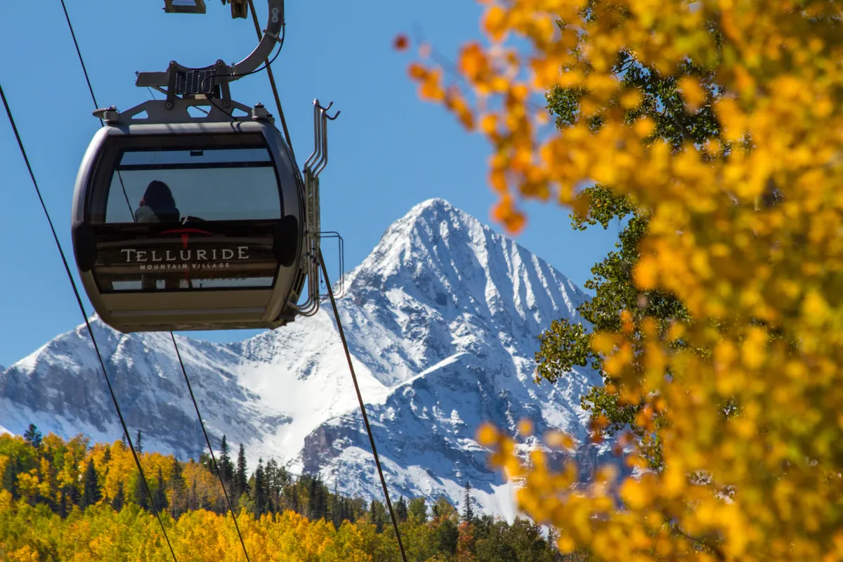 A gondola gliding over tree-lined slopes with vibrant autumn foliage and snowy peaks in the background. The scene is a dynamic mix of Telluride’s signature outdoor activities and accessibility, evoking adventure and easy access to town attractions.