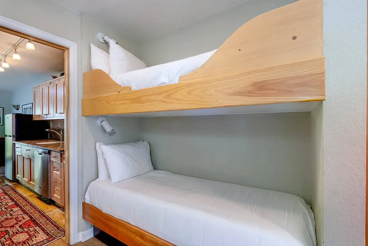 A view of the bunk nook with well-made bunk beds adjacent to the kitchen entrance and a textured rug, conveying a sense of warmth and togetherness. The open layout allows the bunk beds to feel connected to the main living space, reinforcing the family-friendly, communal environment described.