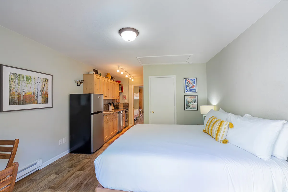 An open-plan space that includes a kitchenette with stainless steel appliances adjacent to a cozy bed with white linens. Artwork on the walls and a wooden dining chair add touches of comfort and personality. The kitchen area’s proximity to living and sleeping spaces is clear, emphasizing easy movement between areas.
