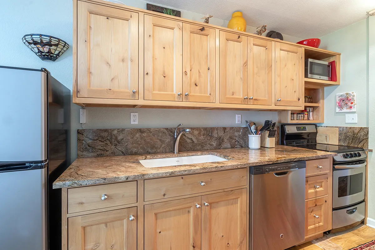 A warm, inviting kitchen featuring wooden cabinets, a sleek granite countertop, and stainless steel appliances. The layout is compact yet functional, with enough space for multiple people to participate in meal preparation or socializing. A sink and ample surface area support a variety of cooking activities.
