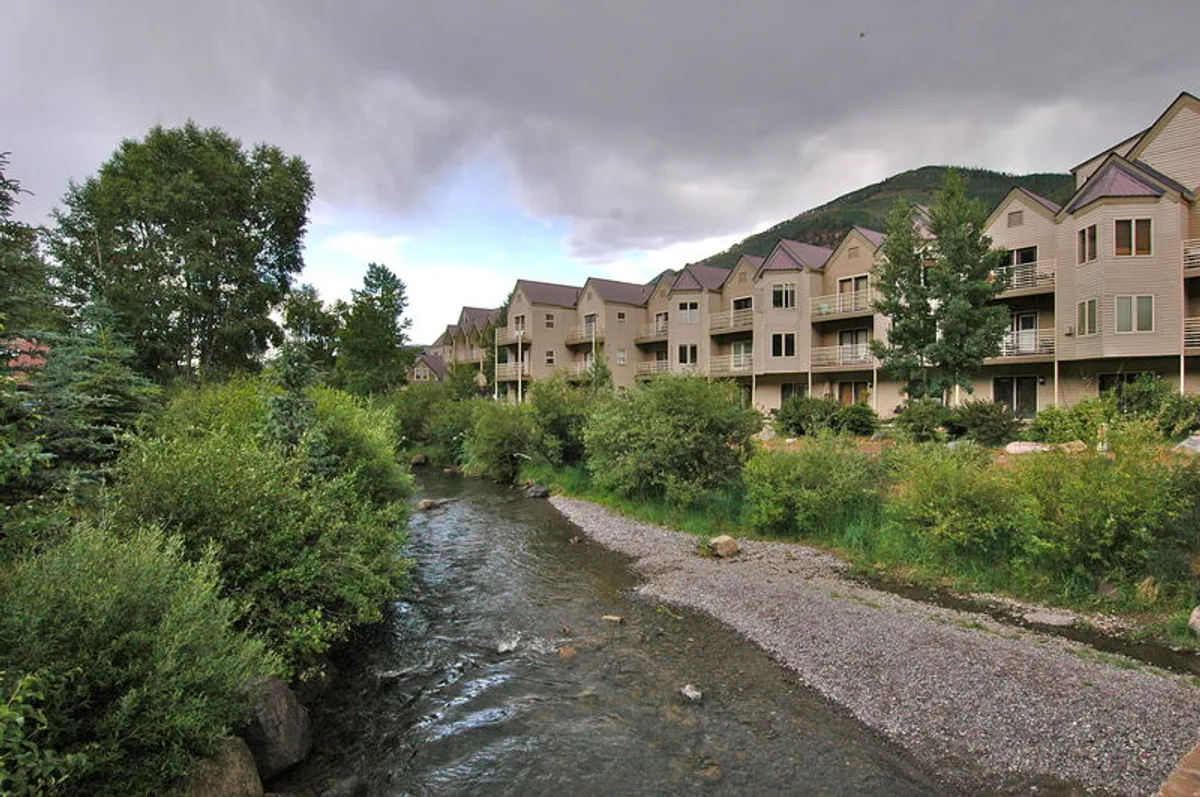 A gentle stream runs alongside residential buildings, framed by leafy trees. The scene feels serene and accessible, suggesting immediate access to natural attractions like the river trail for walking, biking, or peaceful moments by the water.