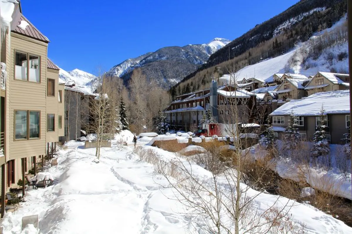 This image captures a tranquil winter scene with snow-covered ground, a mountain backdrop, wooden buildings, and snow-covered trees. The area is clearly set up for outdoor winter activities, suggesting direct access to skiing, snowboarding, and nearby trails.